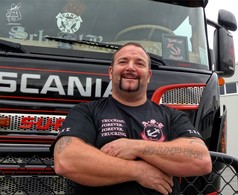 Ein Mann tr�gt ein schwarzes AST Fan Shirt aus Baumwolle/Viskose mit A-S-C Anker-Logo und "TRUCKING. FOREVER." Aufdruck. LKW im Hintergrund.